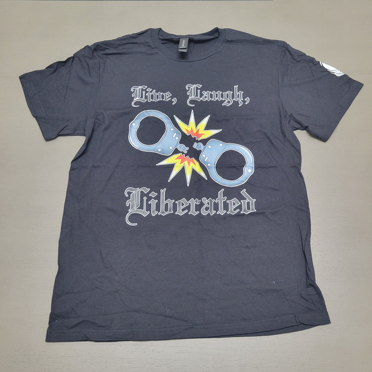 A black short-sleeve t-shirt laid flat on a neutral gray background. The shirt features a graphic print with the gothic text 'Time, Laugh, Liberated' surrounding a graphic of handcuffs with a yellow and orange starburst explosion in the center.
