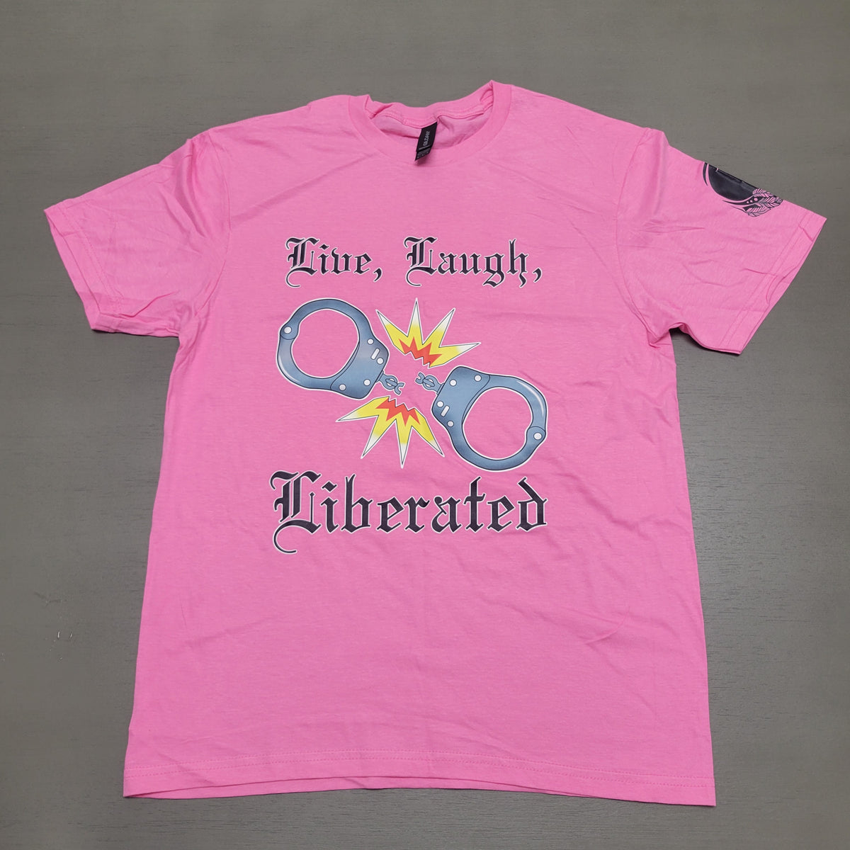 A bright pink short-sleeve t-shirt laid flat on a gray surface. The front features a graphic of handcuffs bursting from a yellow and orange explosion, with the gothic text 'Live, Laugh, Liberated' printed in black.