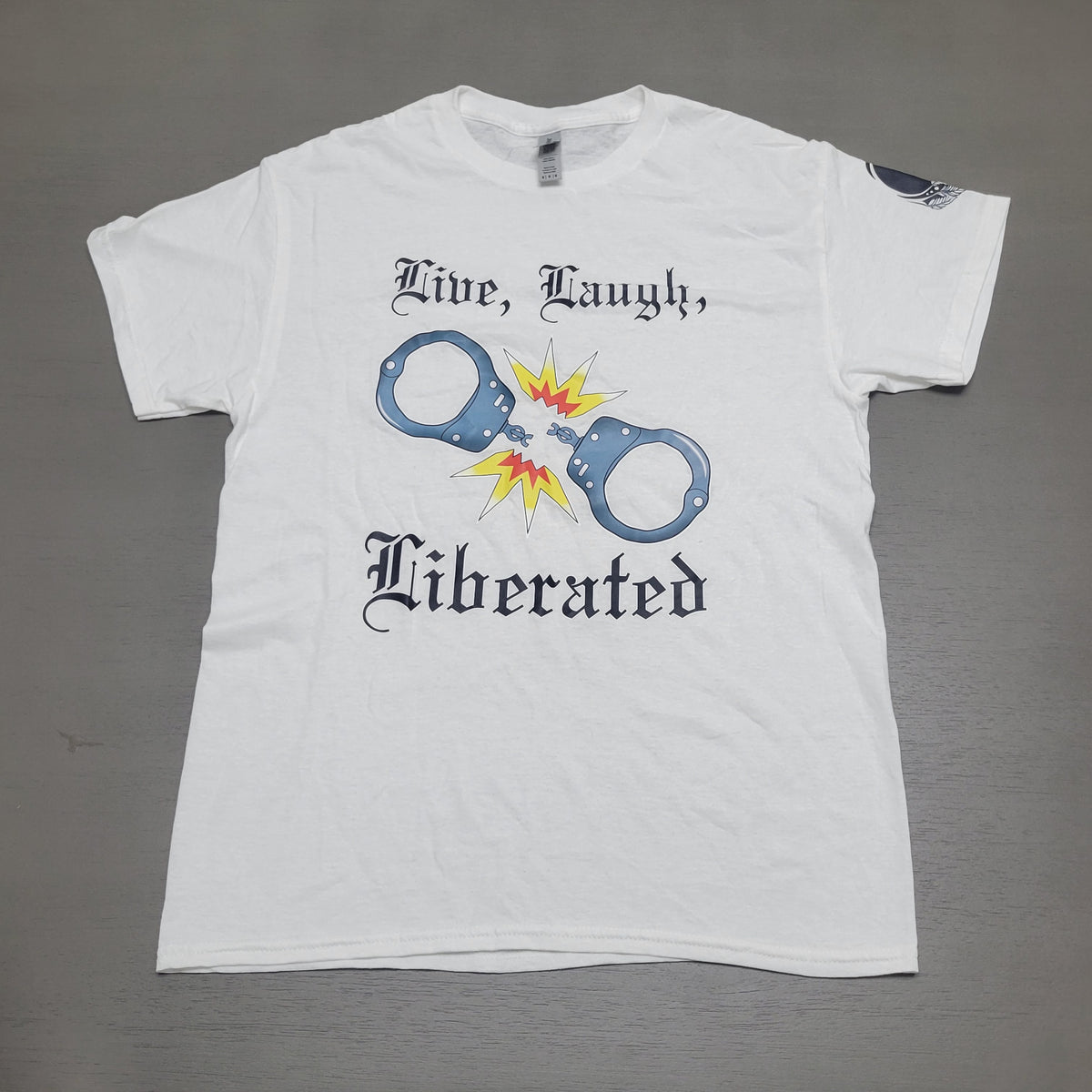 A white short-sleeve graphic t-shirt laid flat on a gray surface. The shirt features a graphic of handcuffs with a yellow and orange explosion in the center, flanked by the black gothic text 'Mine, Laugh,' and 'Liberated'.